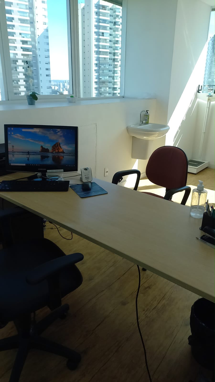 Sala do consultório com mesa, computador, cadeiras e janelas amplas